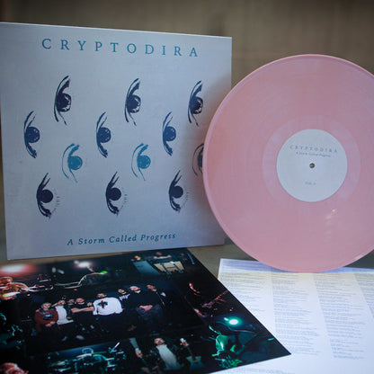 A limited edition pale pink Cryptodira vinyl sits beside its album cover—featuring stylized blue eyes and "Cryptodira: A Storm Called Progress"—with an insert of band photos and a lyric sheet placed below.