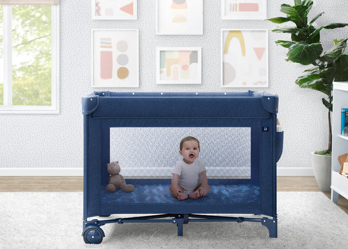 Baby sitting inside a blue crib in a room with a teddy bear and framed pictures on the wall.
