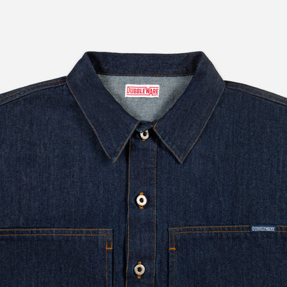 Close-up of a dark indigo denim button-up shirt by Dubble Wade, showing the collar, front buttons, chest pockets, and a small brand tag on the right chest pocket.