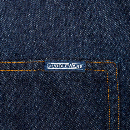 Close-up of dark indigo denim fabric with orange contrast stitching and a small rectangular Dubbleware label near the pocket.