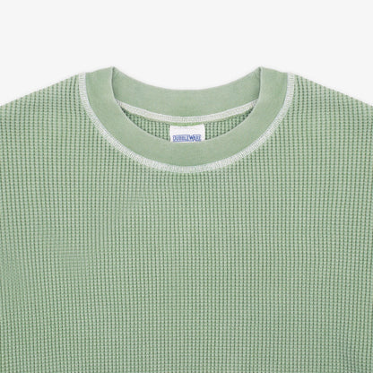 Close-up of a light sage green waffle-knit crewneck sweater with a white ribbed collar trim and a visible neck label reading 'SWEATWEAR'.
