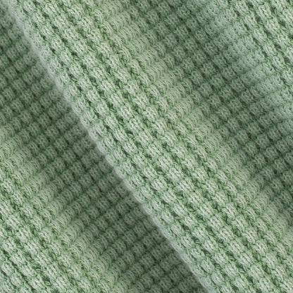 A close-up of a light sage green knit fabric with a textured, ribbed pattern and soft diagonal folds.