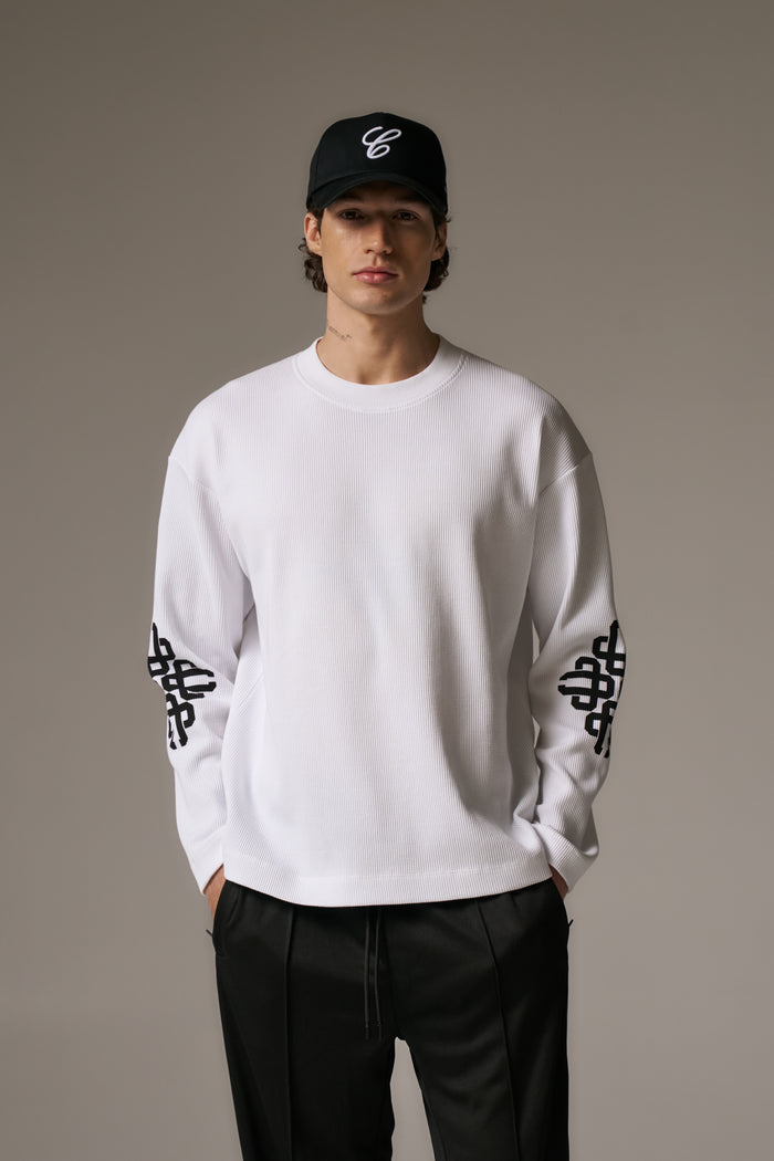Person wearing a white long sleeved t-shirt with black patterns and a black cap on a grey background