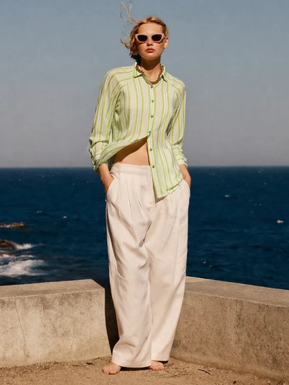 Person wearing a light green striped shirt and white pants standing by the ocean.