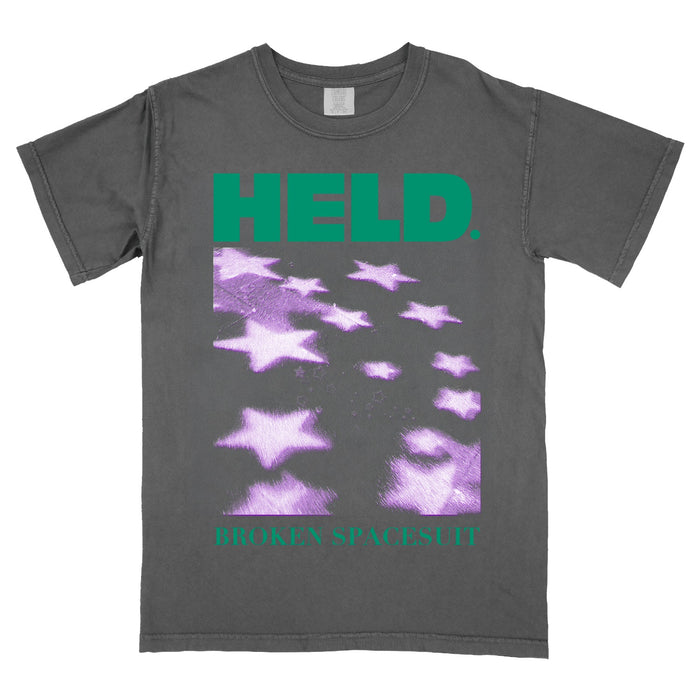 Gray Comfort Colors t-shirt with green text reading HELD. at the top and BROKEN SPACESUIT at the bottom. The center features blurred, purple star shapes scattered diagonally—a unique choice for fans of printed shirts.