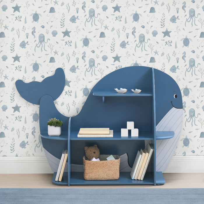 A blue whale-shaped bookcase with seven shelves, filled with books and decorative items, positioned in a room with wall paper featuring sea creatures.